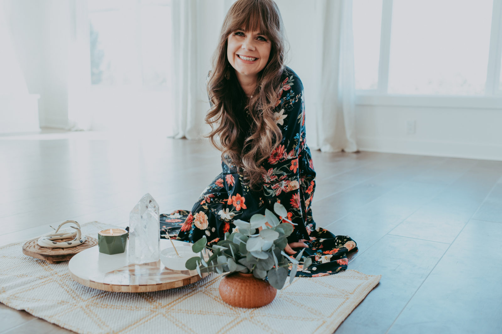 Woman (Briana) in a dark floral dress smiling while seated on a floor mat with a crystal point, candle, antlers, and eucalyptus arrangement on a tray in a light-filled room.