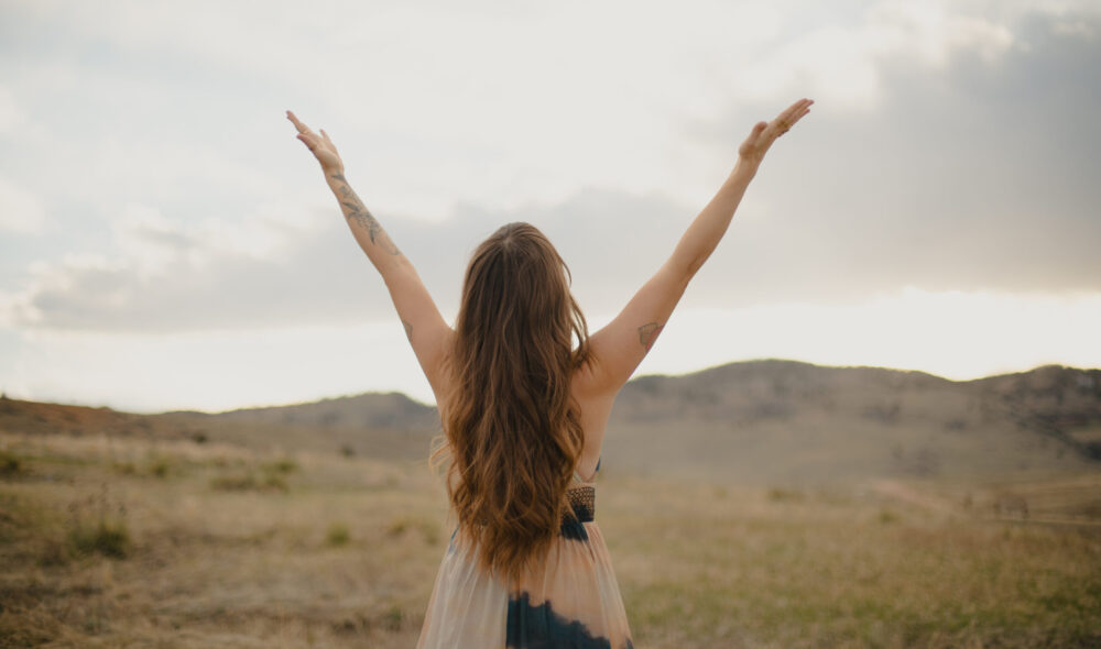 Woman (Briana Borten) with long hair raising arms toward the sky in an open field at sunset, symbolizing freedom and spiritual connection.