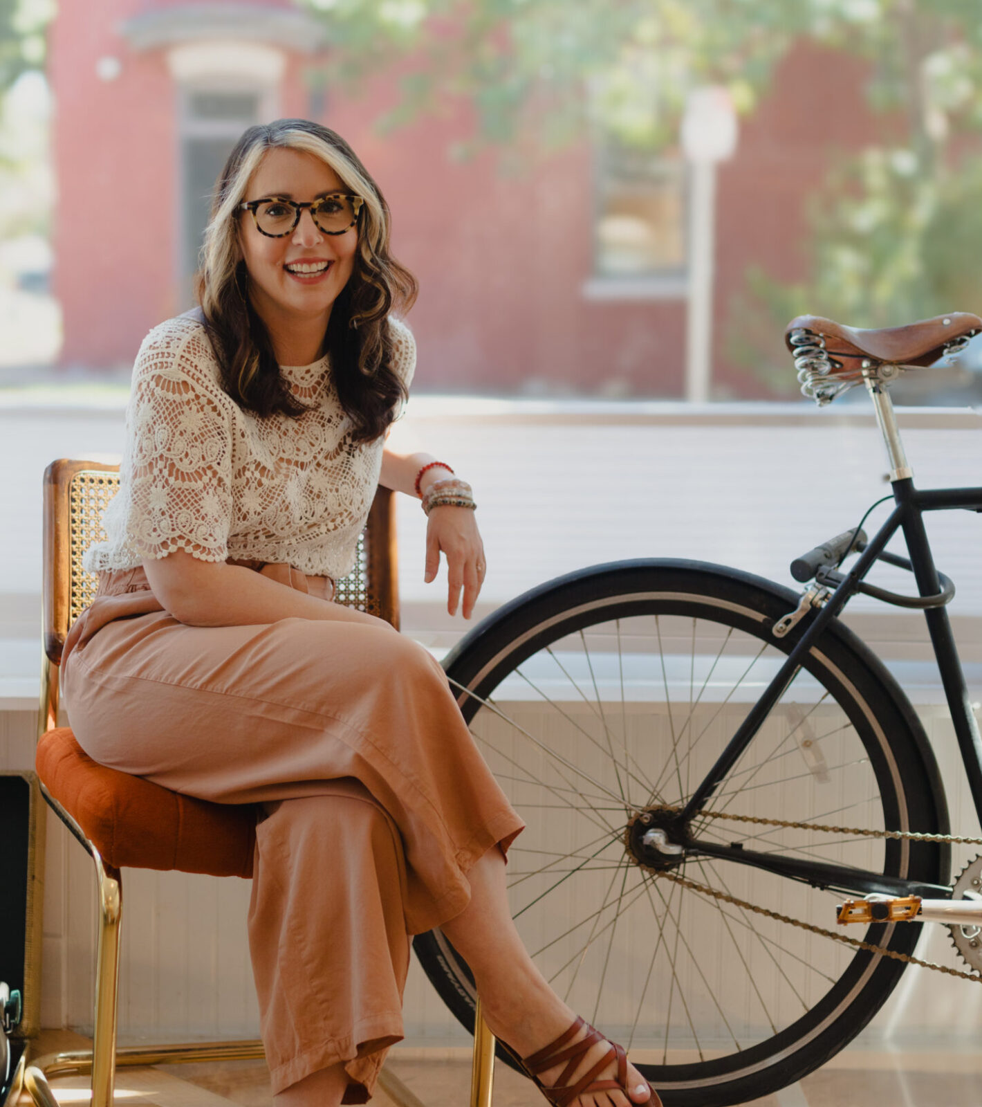 Alyssa smiling while sitting on a chair indoors next to a bicycle, wearing a white lace top and light brown pants.