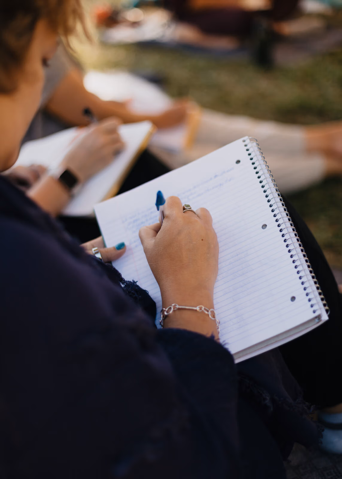 Person writing in a spiral notebook during an outdoor gathering or workshop.