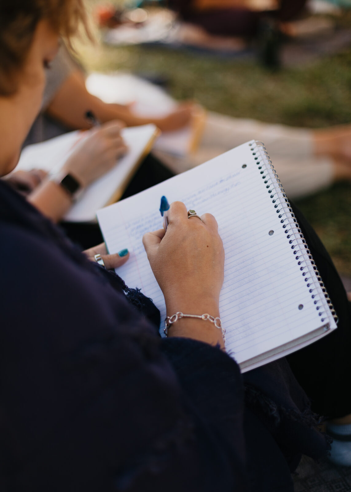 Person writing in a spiral notebook during an outdoor gathering or workshop.