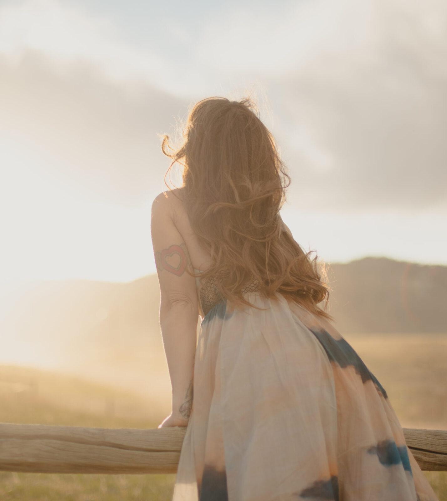 Woman (Briana Borten) with long flowing hair gazes into the glowing sunset over an open field, leaning gently against a wooden fence.