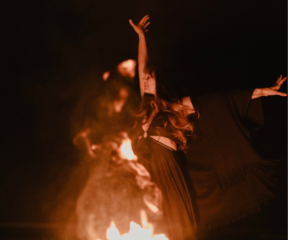 A woman (Briana Borten) in flowing dark clothing dances with her arms raised beside a glowing fire at night